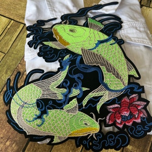 May include: Embroidered patch featuring two green koi fish swimming in a black and blue water design with a red flower. The patch is surrounded by a black border with a blue and white design.