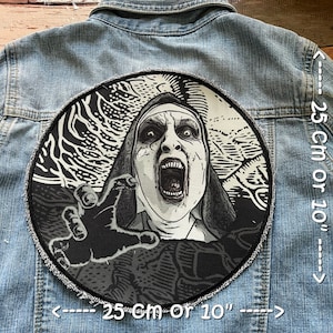 May include: Black and white embroidered patch of a nun with a menacing expression, her hand reaching out towards the viewer. The patch is 25 cm in diameter and is sewn onto a denim jacket.