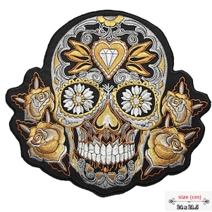 Op de afbeelding: Geborduurde strijkpatch met een zwart-witte suikerschedel met gouden accenten. De schedel heeft een diamant in een hartvorm op zijn voorhoofd, bloemmotieven rond de ogen en rozen aan de zijkanten. De patch is 24 x 21,5 cm.