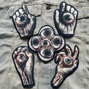 May include: Five iron-on patches featuring a hand holding an eyeball. The patches are black with a white and red design. The patches are perfect for adding a spooky touch to clothing or bags.