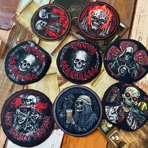 May include: Eight embroidered patches with various skull and skeleton designs. The patches are circular and feature black, white, red, and gray colors. The patches are on a wooden surface.