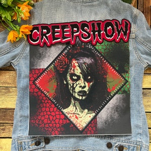 May include: A denim jacket with a red and white embroidered patch that says "CREEP SHOW". The patch is above a diamond-shaped patch with a graphic of a zombie woman with blood splatters on her face and a rose in her hair.