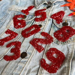 May include: A denim jacket with red rhinestone numbers 1, 5, 4, 7, 0, 2, 3, 8, and 9 sewn on the front.