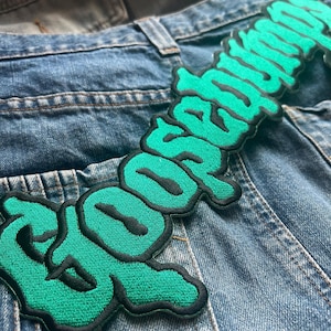 May include: A green and black embroidered patch with the word "Goosebumps" in a stylised font. The patch is sewn onto a pair of blue denim jeans.