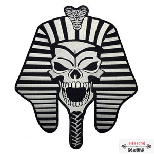 May include: A black and white embroidered patch featuring a skull with an open mouth, wearing a pharaoh headdress. The patch includes a cobra at the top. The patch measures 24 x 27.5 cm.