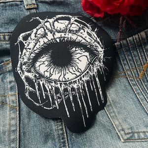 May include: Black and white embroidered patch of a dripping eye with a detailed design. The eye is surrounded by a black background and white dripping lines.