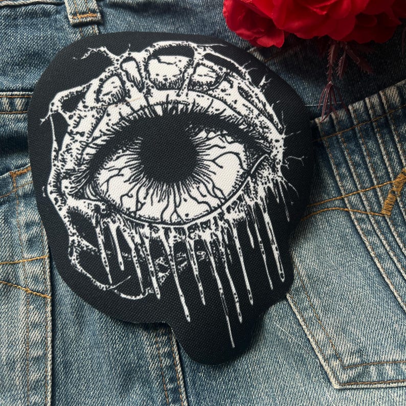 May include: Black and white embroidered patch of a dripping eye with a detailed design. The eye is surrounded by a black background and white dripping lines.