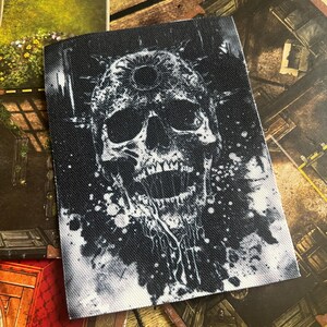 May include: A black and white skull with a third eye on its forehead, surrounded by white paint splatters on a black background.