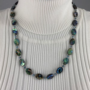 May include: Abalone shell necklace with oval-shaped iridescent beads in shades of blue, green, and brown. The beads are strung together with small blue beads. The necklace is displayed on a white turtleneck.