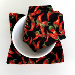 May include: A white ceramic bowl rests on a black square placemat and a matching square napkin. Both the placemat and napkin feature a repeating pattern of red chili peppers with green stems and leaves. The background is white.