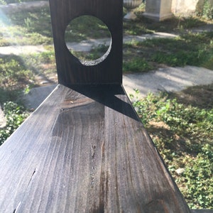 May include: Close-up of a dark stained wooden structure with a circular hole. The wood grain is visible, and the structure has a triangular shape. The background shows a glimpse of a garden.