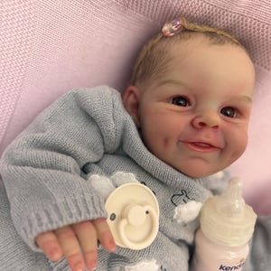Puede incluir: Una mu&ntilde;eca beb&eacute; realista con una expresi&oacute;n sonriente, vestida con un su&eacute;ter azul claro y un peque&ntilde;o accesorio para el cabello. La mu&ntilde;eca tiene un chupete y un biber&oacute;n. La mu&ntilde;eca est&aacute; sobre una superficie rosa.