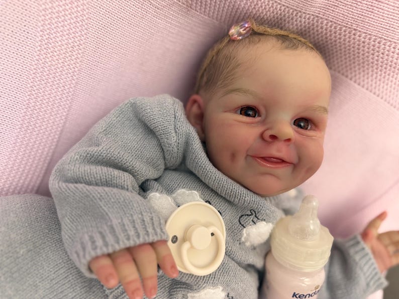 Puede incluir: Una mu&ntilde;eca beb&eacute; realista con una expresi&oacute;n sonriente, vestida con un su&eacute;ter azul claro y un peque&ntilde;o accesorio para el cabello. La mu&ntilde;eca tiene un chupete y un biber&oacute;n. La mu&ntilde;eca est&aacute; sobre una superficie rosa.