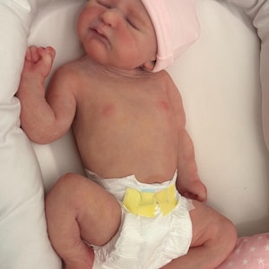 May include: A realistic baby doll with closed eyes, wearing a pink cap and a diaper with a yellow bow. The doll is lying in a white cushioned nest, with a pink pillow with white stars in the background.