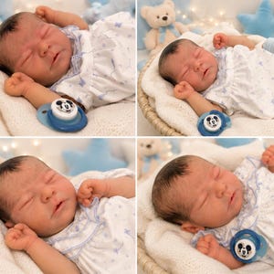 May include: A realistic newborn baby doll with dark hair, sleeping in a white dress with a blue butterfly pattern. The doll has light skin, closed eyes, and is accompanied by a blue pacifier featuring a Mickey Mouse graphic on the front.