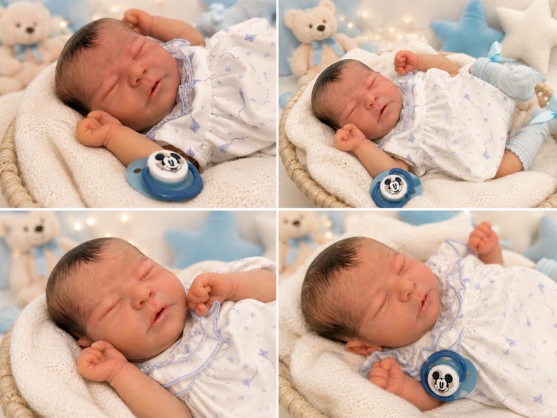 May include: A realistic newborn baby doll with dark hair, sleeping in a white dress with a blue butterfly pattern. The doll has light skin, closed eyes, and is accompanied by a blue pacifier featuring a Mickey Mouse graphic on the front.
