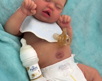 May include: A realistic newborn baby doll with light skin, closed eyes, and brown hair. The doll wears a white bib with a scalloped edge, a white diaper, and a translucent pacifier. A small white baby bottle with a NAN 1 label sits nearby.