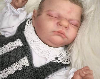 May include: A sleeping baby doll wearing a gray knitted vest with white lace trim and a white shirt with lace trim.