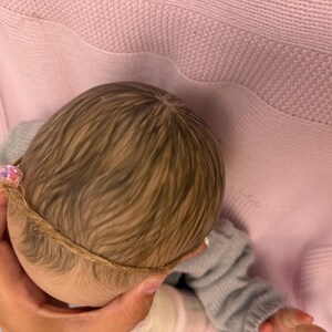 Puede incluir: Cabeza de mu&ntilde;eca con cabello casta&ntilde;o claro y una delicada diadema. La mu&ntilde;eca lleva un su&eacute;ter gris y descansa sobre una manta rosa. El cabello de la mu&ntilde;eca est&aacute; peinado con una sutil onda.