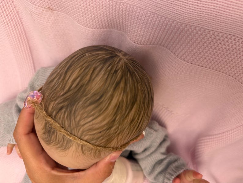 Puede incluir: Cabeza de mu&ntilde;eca con cabello casta&ntilde;o claro y una delicada diadema. La mu&ntilde;eca lleva un su&eacute;ter gris y descansa sobre una manta rosa. El cabello de la mu&ntilde;eca est&aacute; peinado con una sutil onda.