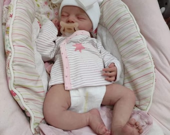 Puede incluir: Una muñeca bebé durmiendo que lleva un gorro blanco con un lazo, una camisa a rayas rosa y blanca y un pañal. La muñeca está tumbada en una hamaca para bebés con estampado rosa y blanco.
