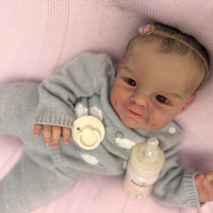 Puede incluir: Una mu&ntilde;eca beb&eacute; realista con un conjunto de punto gris, un chupete y un biber&oacute;n. La mu&ntilde;eca tiene tez clara, ojos marrones y un peque&ntilde;o lazo rosa en el pelo. La mu&ntilde;eca est&aacute; tumbada sobre una manta rosa.
