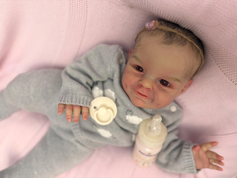 Puede incluir: Una mu&ntilde;eca beb&eacute; realista con un conjunto de punto gris, un chupete y un biber&oacute;n. La mu&ntilde;eca tiene tez clara, ojos marrones y un peque&ntilde;o lazo rosa en el pelo. La mu&ntilde;eca est&aacute; tumbada sobre una manta rosa.