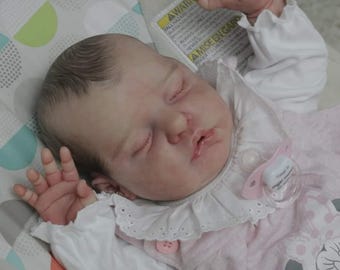 May include: A sleeping baby doll with brown hair, wearing a pink and white dress with a white lace collar and a pink pacifier. The doll is lying on a white and green patterned blanket.