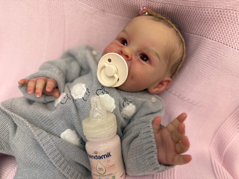 Puede incluir: Una mu&ntilde;eca beb&eacute; realista con un su&eacute;ter azul claro con dise&ntilde;os de nubes blancas. La mu&ntilde;eca tiene un chupete en la boca y un biber&oacute;n de f&oacute;rmula Kendamil. La mu&ntilde;eca est&aacute; acostada sobre una manta rosa.