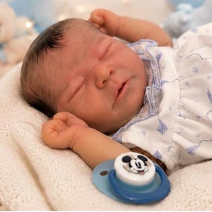 May include: A realistic newborn baby doll with dark hair and closed eyes, wearing a white short-sleeved shirt with a blue kite pattern. The doll is lying down with arms raised and has a blue pacifier featuring a Mickey Mouse graphic nearby.
