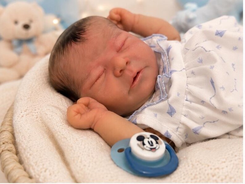 May include: A realistic newborn baby doll with dark hair and closed eyes, wearing a white short-sleeved shirt with a blue kite pattern. The doll is lying down with arms raised and has a blue pacifier featuring a Mickey Mouse graphic nearby.