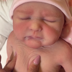 May include: Close-up of a newborn baby with closed eyes, wearing a light pink beanie. The baby's skin is pink with red cheeks. A hand gently touches the baby's chest.
