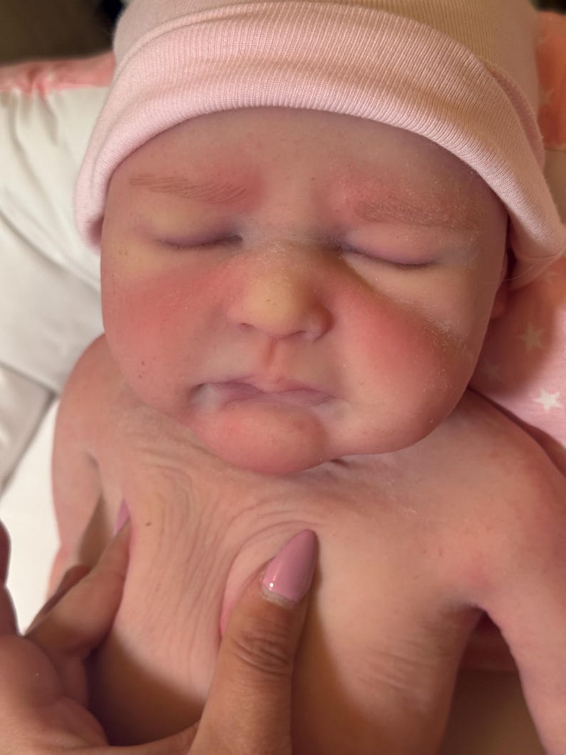 May include: Close-up of a newborn baby with closed eyes, wearing a light pink beanie. The baby's skin is pink with red cheeks. A hand gently touches the baby's chest.