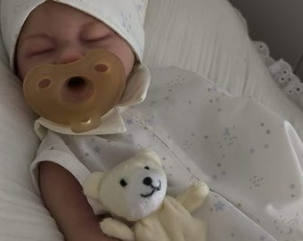 May include: A white baby doll with a dummy in its mouth, wearing a white and blue star patterned dress and a white hat with blue stars. The doll is holding a white teddy bear.