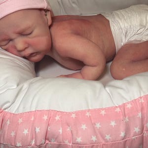 May include: A realistic baby doll wearing a pink knit cap and a diaper, lying on a white pillow inside a pink and white star-patterned bed. The doll has a realistic skin tone and is posed as if sleeping.