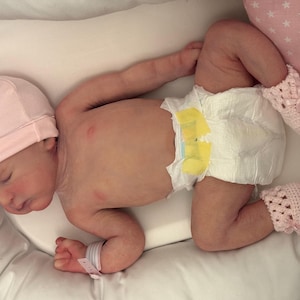 May include: A newborn baby wearing a pink knit cap, a diaper, and pink crocheted booties, resting on a white pillow. The baby has a hospital wristband. A pink star-patterned pillow is in the background.
