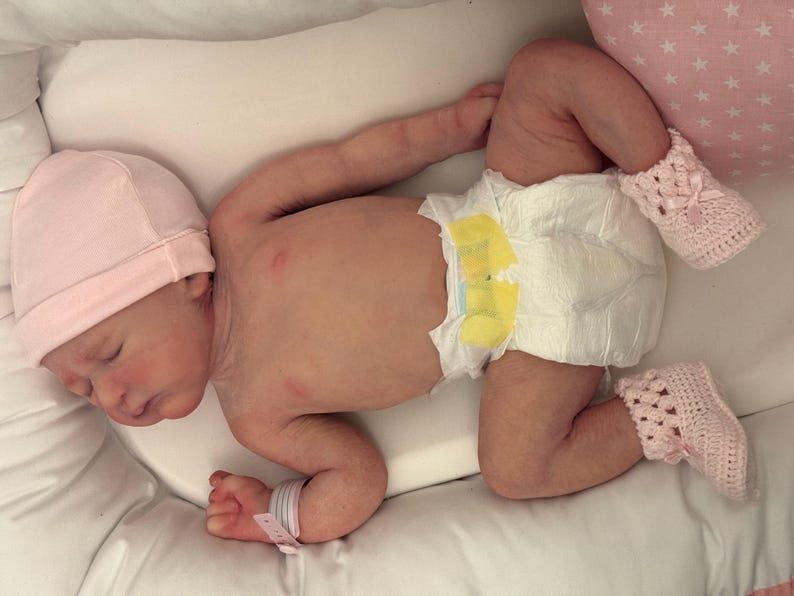 May include: A newborn baby wearing a pink knit cap, a diaper, and pink crocheted booties, resting on a white pillow. The baby has a hospital wristband. A pink star-patterned pillow is in the background.