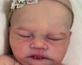 Puede incluir: Un muñeco de bebé durmiendo con una diadema de flores rosa y blanca. El muñeco tiene rasgos realistas y está hecho de silicona suave.