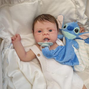 Può includere: Una bambola realistica di un bambino con capelli castani e occhi azzurri indossa una tutina bianca e un ciuccio blu. La bambola è sdraiata su una coperta bianca con una coperta a righe blu e bianche e un giocattolo di peluche blu.