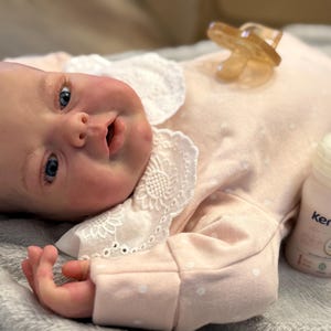 Może przedstawiać: Lalka niemowlęca w różowym pajacyku w białe grochy i białym koronkowym kołnierzyku. Smoczek i butelka mleka modyfikowanego Kendamil leżą obok lalki. Dziecko ma niebieskie oczy i różowe policzki.