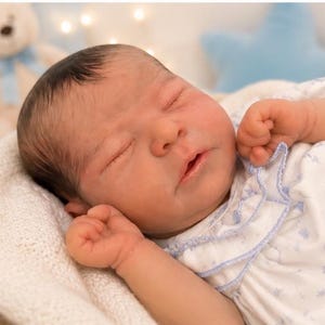 May include: A newborn baby doll with dark hair and closed eyes, wearing a white short-sleeved shirt featuring a light blue kite pattern and a ruffled collar. The doll is resting in a white textured basket with its hands positioned near its face.