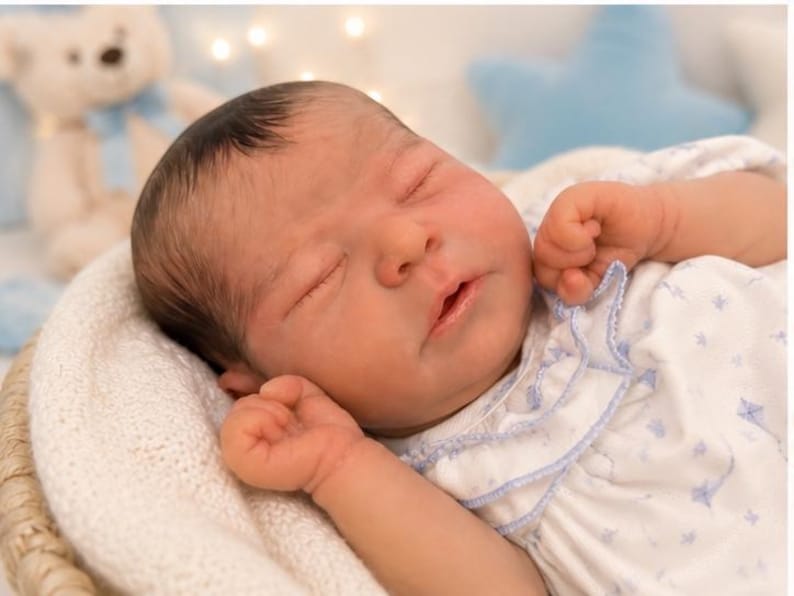 May include: A newborn baby doll with dark hair and closed eyes, wearing a white short-sleeved shirt featuring a light blue kite pattern and a ruffled collar. The doll is resting in a white textured basket with its hands positioned near its face.