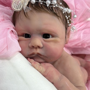 May include: A lifelike baby doll with dark hair and large, dark eyes. The doll wears a pearl and silver headband and a delicate butterfly hair accessory. The doll is lying on a white surface, with a pink tulle background.