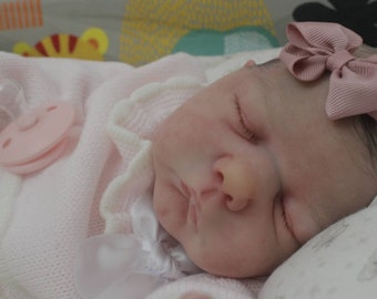May include: A sleeping baby doll with closed eyes, wearing a pink knitted outfit with white lace trim and a pink bow headband. The doll is lying on a white blanket with a pink pacifier.