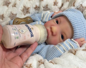 May include: A baby doll in a blue knitted hat and sweater is being fed from a pink and white Kendamil baby bottle. The bottle has a white teat and is held by a hand. A dummy is in the background.