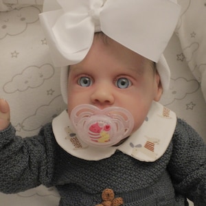 Puede incluir: Una muñeca bebé con ojos azules y un lazo blanco en la cabeza. La muñeca lleva un suéter de punto gris y un babero blanco con un patrón marrón y blanco. La muñeca sostiene un chupete rosa y blanco en su boca.