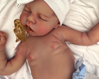 Puede incluir: Una muñeca bebé realista con los ojos cerrados, que lleva un gorro blanco de punto y un pañal azul y blanco. La muñeca tiene un chupete en la boca y está tumbada sobre una manta blanca con dibujos de estrellas rosas.
