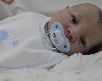 May include: A baby doll with blue eyes is lying on a white blanket. The doll is wearing a white onesie with blue embroidery and a blue pacifier.