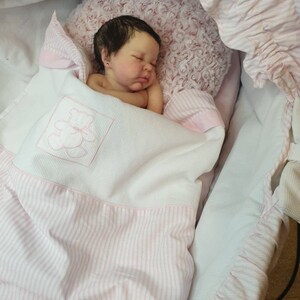Pu&ograve; includere: Un bambolotto che dorme con i capelli scuri e gli occhi chiusi, sdraiato su una coperta a righe rosa e bianche in una culla di vimini bianca.