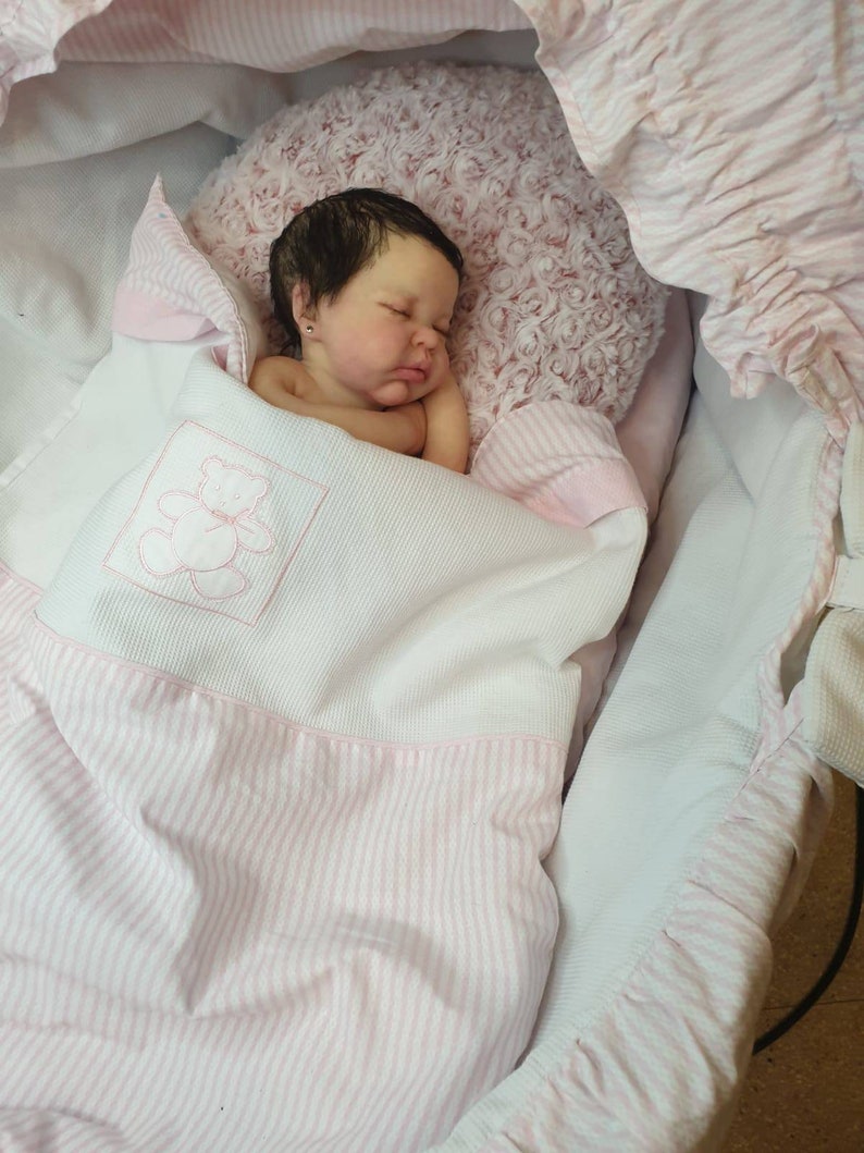 Pu&ograve; includere: Un bambolotto che dorme con i capelli scuri e gli occhi chiusi, sdraiato su una coperta a righe rosa e bianche in una culla di vimini bianca.